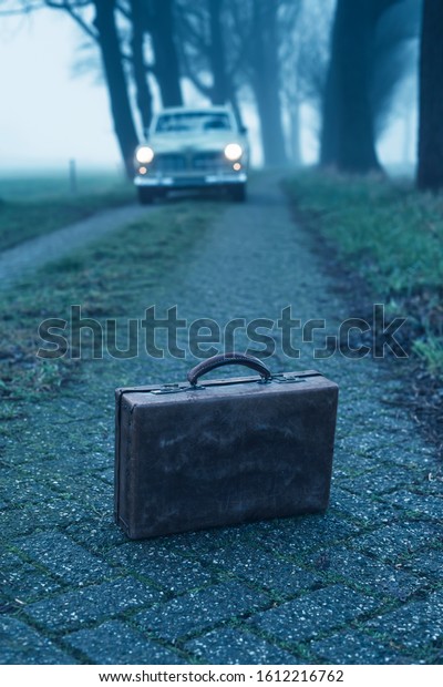 Old briefcase on country road during foggy\
dusk with classic car in the\
background.