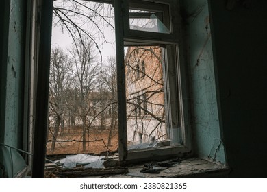 Old broken metal frame window sill of shabby dirty painted walls with black destroyed spider nets and overlooking at green fall trees brick wall building under cloudless blue sky in daytime
