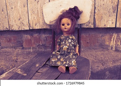 Old Broken Soviet Doll In A Dress On A Broken Chair Near A Shabby House. Darkly Tinted Photo.