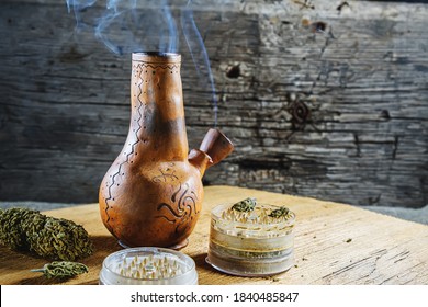 Old brown steamung bong and open cannabis grinder with chopped marijuana weed and medical cannabis in smoke by wooden desck and sackcloth. Horizontal