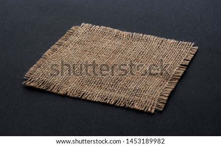 Old burlap fabric napkin closeup. Rough linen jute, sackcloth piece isolated on black background. Hessian texture cloth tag with frayed edges. Dark sack material