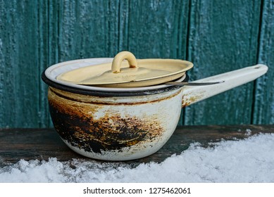 An Old Burnt Saucepan Covered By A Lid Stands On A Wooden Table In White Snow Against A Green Wall In The Street