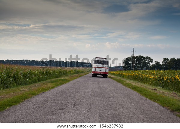 Old bus carrying passengers on rural road in\
between corn and sunflower\
fields