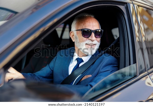 An old businessman drives a car. A happy senior\
businessman sits in his car with his hands on the steering wheel\
and looks through the\
window.