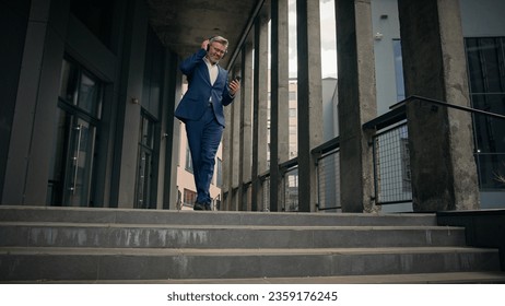 Old businessman middle-aged Caucasian employee senior business man worker listening to music in headphones holding mobile phone dancing walking down stairs office in city happy good mood success jump - Powered by Shutterstock