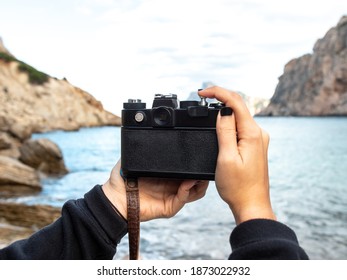 An old camera held by two women's hands on a rocky beach by the sea