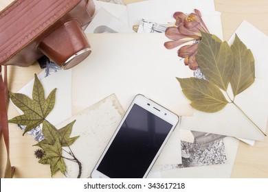 Old camera and smartphone on stack of photos with dry plants, on wooden surface.