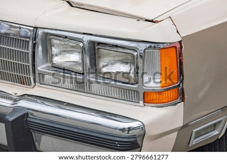 Old car front section, featuring classic rectangular headlights and worn white bumper showcasing rusted elements and aged chrome trim detailing against an out-of-focus background emphasizing well-used
