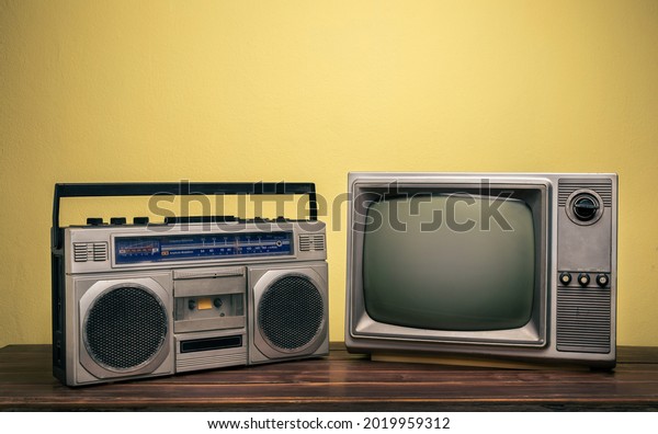 An old cassette radio and vintage\
TV on wooden table in front of yellow concrete\
wall.