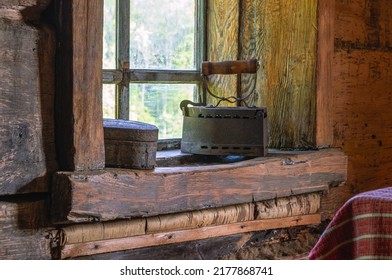 An Old Cast-iron Coal Iron Made In The 19th Century On The Windowsill Of An Old Wooden House. The Interior Of An Old House. Dark Log Walls Of The House. Rustic Architecture.