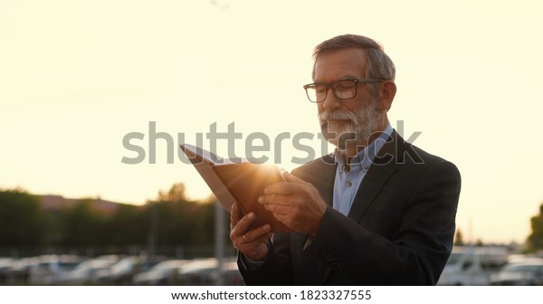 Old\
Caucasian gray-haired man in glasses walking and reading book\
outdoor in sunset at cars parking on background. Senior male\
professor strolling and read textbook.\
Studying.
