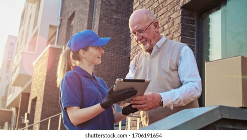 Old Caucasian male pensioner standing outdoors at his house and taking parcel from pretty young female postal worker courier. Woman delivering box to pensioner. Man signing. Mailing service.