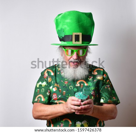 Old Caucasian man with a long white beard, festive St Patrick's Day shirt, glasses with the Irish colors and a green drink making faces and showing emotions.

