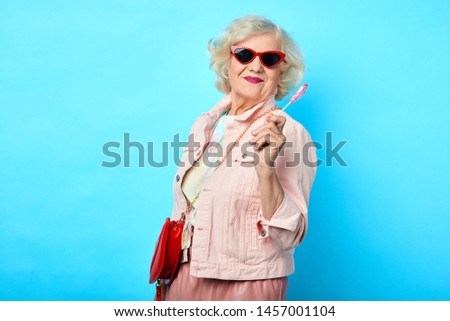 Old cheerful happy lady with sunglasses holding lollipop and looking at the camera on a blue background. sweets, love, valentine's day. fashion concept
