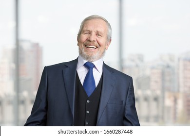 Old cheerful male manager laughing out loud. Portrait of senior boss in suit. Blurred windows with view on the city background.