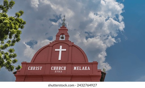 The old Christ Church. The upper part of the building is painted red. Dome, cross, bell on a background of blue sky and clouds.  White lettering with the name and year of construction. Malacca. 