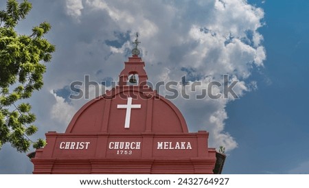 The old Christ Church. The upper part of the building is painted red. Dome, cross, bell on a background of blue sky and clouds.  White lettering with the name and year of construction. Malacca. 