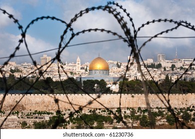 The Old City of Jerusalem, including the Dome of the Rock and various church steeples, seen through coils of razor wire, illustrating the Holy Land's history of division and conflict.