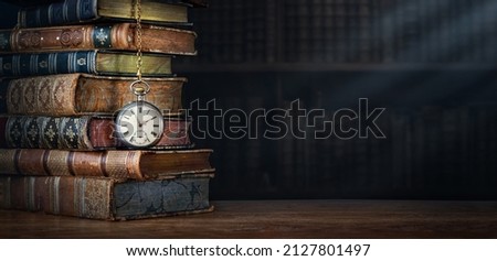 Old clock hanging on a chain on background of old books. Retro clock as a symbol of time a books are a symbol of knowledge. Concept on the theme of history, nostalgia, culture, vintage,  antique. 