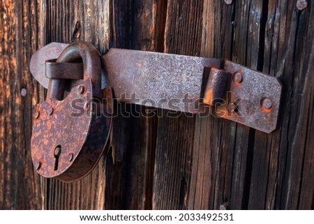 An old closed rusty barn lock hanging on the shackle of a wooden door. View from the right. Close-up. Soft light.