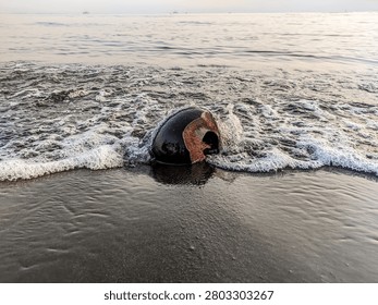 An old coconut shell hit by a gentle incoming sea wave and white foam on a wet black sand beach.