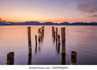 Old columns of abandoned pier standing in calm sea coast water in the morning sunrise orange light with hills, smoke and clouds in the background - central symmetrical perspective composition - Powered by Shutterstock