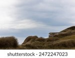 An old concrete bunker sits atop a grassy coastal hill, set against an overcast sky with dramatic clouds overhead