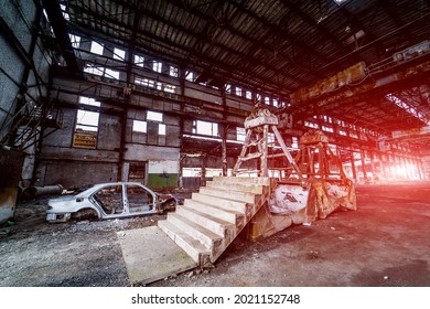 Old Construction With Stairs And Antique Car Frame On The Background Of Broken Plant. Interior Of The Large Abandoned Building With A Damaged Car And Metal Debris