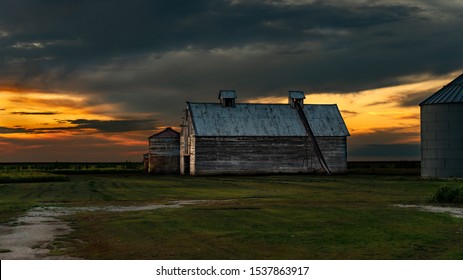 Old Corn Crib Or Barn And Metal Grain Bins Of A Family Farm In The Midwest With A Colorful Orange Sunset / Sunrise & Storm Clouds Threatening. Concepts Of Family Farms, Trade War, Tariffs. 