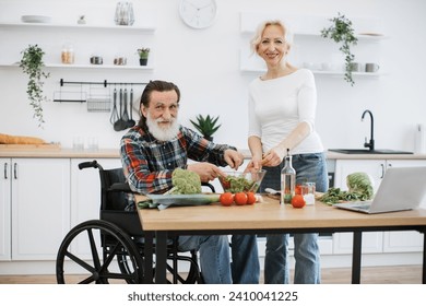 Old couple with disability spends free time cooking breakfast in modern light kitchen looking at camera. Senior husband in wheelchair and wife mixing chopped vegetables in bowl while prepare salad. - Powered by Shutterstock