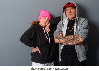 Old Couple Dressed In Hip-hop Style. Stylish Grandma Leant On Her Husband. Horizontal Photo With Gray Background.