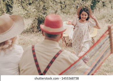 Old Couple In The Garden Sitting On Hammock Back View. Grandfather In Red Hat Loves Grandmother. Happy Old Age. Young Granddaughter Runs To Them