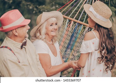 Old Couple In The Garden Sitting On Hammock Back View. Grandfather In Red Hat Loves Grandmother. Happy Old Age. Young Granddaughter Runs To Them