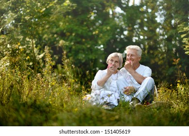 Old Couple Sitting In Grass In Forest And Eating Apples