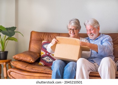 Old Couple Unpacking Delivery Box At Home. Happy Senior Couple Looking At Carton Box While Sitting On Sofa In Living Room At Home. Elderly Couple Checking Out Delivered Stuff At Home.