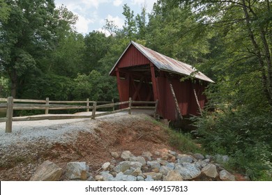An old covered bridge in South Carolina, USA on a hot, humid summer afternoon with a blue sky, white clouds and a forest of green trees. Rocky foreground, wooden rail fence on a horizontal format.