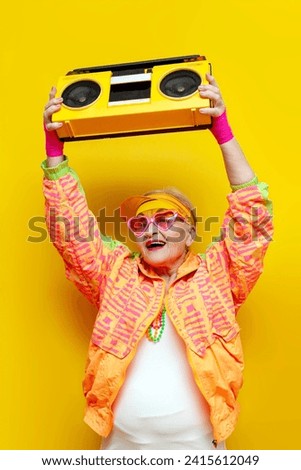 old crazy grandmother in fashionable sportswear listens loudly to music on a tape recorder and dances on a yellow isolated background, an elderly funny pensioner with glasses raises up a boombox at a