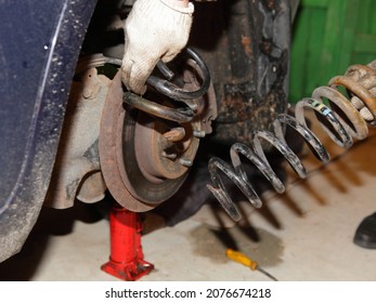 Old Damaged Coil Spring In Hands On Broken Car On Bottle Hydraulic Jack Background - Vehicle Suspension Repair In Garage