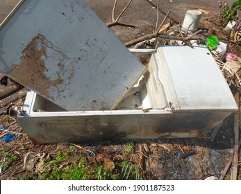 An old, damaged and unusable refrigerator is dumped on the side of the road in a residential park.
