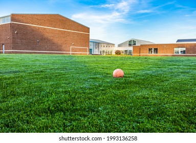 Old And Deflate Orange Football On Green Turf With Sunlight At School Building Background, Depth Of Field. Outdoor Recreation Space For Students. Landscape View Of Empty Soccer Field And Unused Ball.