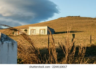 Old dilapidated buildings illuminated by late afternoon sun on side of hill with dark clouds overhead on Central Otago Rail Trail in New Zealand.
