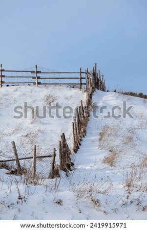 An old dilapidated wooden fence on a farm. The fence line boundary is on a hill enclosing a meadow filled with fresh white snow. Bits of yellow hay sticks out  in the field. There's a blue sky.