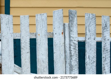 An Old Dilapidated Wooden White Picket Fence With Weathered Boards, Rust Spots, And Rough Posts. There's A Yellow And Green Color House In The Background. The Wall Is Covered With Clapboard Siding. 