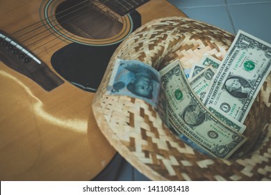 Old Dirty Acoustic Guitar And Multi Currency Money Cash On Weaving Hat. 