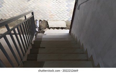 Old Dirty And Damaged Tile Concrete Stairs Going Down Towards Broken Floor Pavement With Rusty Stainless Steel Stair Railing Protection Barrier And White Wall. Closeup Bottom Top View Of Steps.