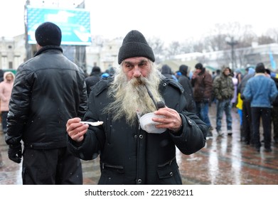 An Old Dirty Homeless Man Standing On Street Smoking Pipe Holding Plastic Plate With Free Food, People Walking Around During Meeting. Revolution Of Dignity. December 7, 2013. Kyiv, Ukraine