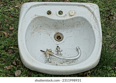 an old dirty sink for hands ceramics with an old broken chrome faucet and two taps lies in the green grass on the street	 - Shutterstock ID 2311998931