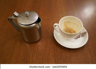 Old Dirty Used Aluminum Tea Pot Kettle With Dirty Used White Tea Mug. Antique Aluminum Kettle With Tea Cup And Empty Used Sugar Sachets. Dirty Coffee And Tea Cup Half Empty On Saucer On Table.