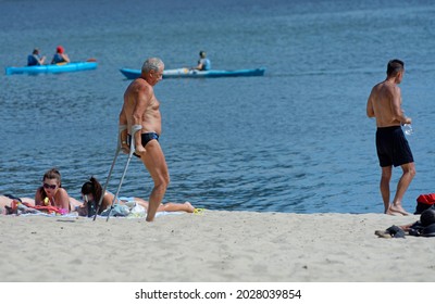 Old Disabled One-legged Man Standing With Double Adjustable Elbow Crutches, People Sunbathing On A River Sandy Beach, Canoes Floating. August 2, 2012. Kyiv, Ukraine