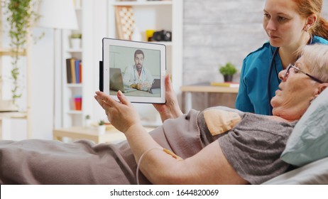 Old disabled woman lying in hospital bed having an online video call with a doctor. A nurse is next to her